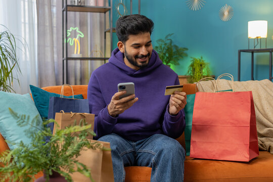 Happy shopaholic consumer indian man sitting with shopping bags at modern home apartment making online order, payment with credit plastic bank card. Discount sale on internet. Young hindu guy on sofa