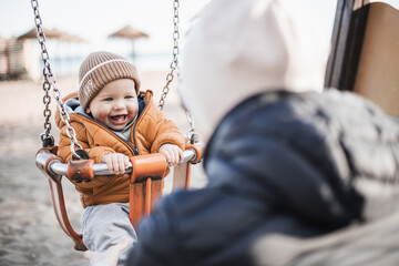 Mother pushing her cheerful infant baby boy child on a swing on sandy beach playground outdoors on...