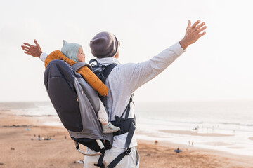 Young father rising hands to the sky while enjoying pure nature carrying his infant baby boy son in backpack on windy sandy beach. Family travel concept
