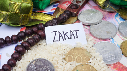 Phrase ZAKAT written on white tag with rice, rosary beads and coins. Selective focus