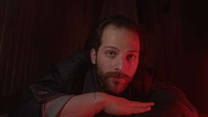 Close-Up Shot of a Young, Long-Haired, Bearded Male Model in a Denim Jacket Striking a Pose and Looking into the Camera under Red and White Lights