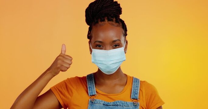 Covid face mask, studio black woman and thumbs up for bacteria safety, corona virus compliance or security policy. Portrait, emoji icon gesture or female with pandemic protection on orange background