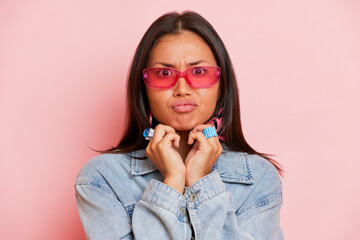 Serious young lady in glasses and denim jacket stands in the middle against a pink background looking at camera with surprise, posing in the studio with hands folded at chin, copy space, high quality