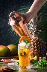 Summer cocktail with vodka, pineapple juice, mango, ice. Long drink or cold mocktail. Bartender shakes copper shaker with splashes, frozen motion and flying drops. Tropical background with palm leaves