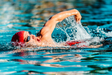 Male swimmer swimming crawl freestyle in blue water outdoor pool. Portrait of an athletic young male triathlete swimming crawl wearing a red cap and swim goggles. Triathlete training for triathlon - obrazy, fototapety, plakaty