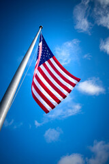 Looking up a metal pole to the American flag waving in the wind against a blue sky with a few puffy, white clouds
