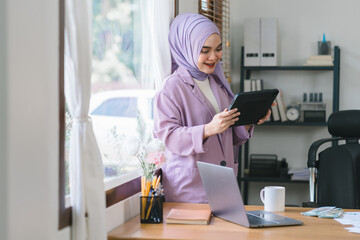 A Muslim millennial businesswoman, wearing a purple hijab, working with tablet and financial report paperwork in home office. The concept of financial advising, teamwork, and accounting is depicted.