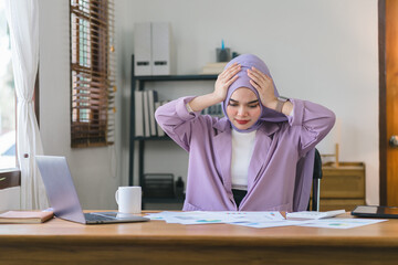 Stressed Muslim millennial businesswoman wearing a purple hijab working from home on laptop looking worried, tired and overwhelmed.