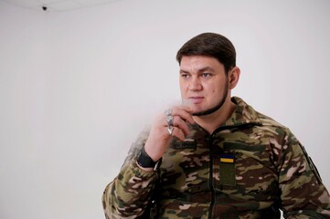 Military man in uniform sitting and smoking He exhales puffs of smoke Ukrainian flag is sewn on protective jacket Adult handsome man