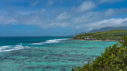 Fototapeta premium The corals are visible through the clear water of the boundless turquoise ocean. The waves are foaming over the reef. The islands with lush tropical vegetation. The villas of the hotel on the shore. 