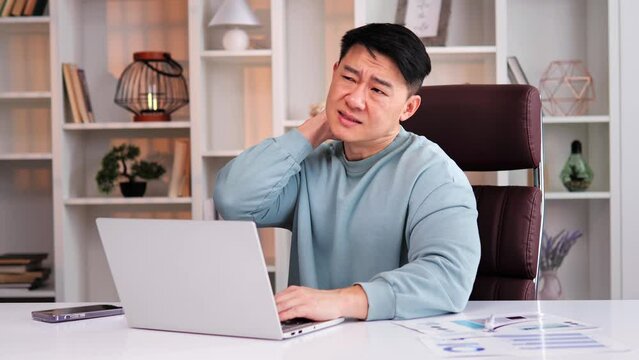 Sudden sharp pain in the neck of Asian office worker. Korean businessman is sitting at desk, massaging neck due to sedentary lifestyle working on computer. Man is trying to relieve tension in muscles.