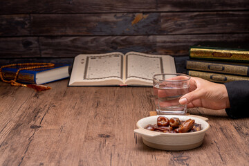 Many Date Palms are on bowls, plates, and cups of water, and the Quran is on wooden tables, to wait for the time of Iftar, with the concept of ramadan Muslims, Arabic calligraphy meaning of Al Quran