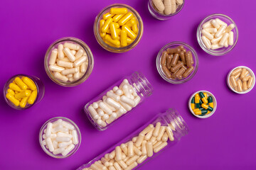 Many small glass bottles and bowls filled with different vitamins and minerals in form of capsules, tablets and pills from above on violet background. Vitamin b, d3, c, a, e, magnesium, zinc.
