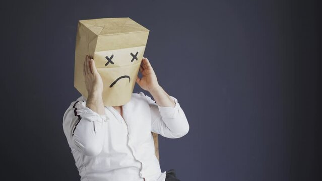 A sad man in a white shirt with a bag on his head, with a sad emoticon drawn, holds his head, closes his eyes in fear. Emotions and gestures.