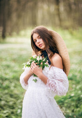 Portrait of a beautiful young brunette in a spring forest. A girl with a bouquet of primroses in her hands in a white lace dress