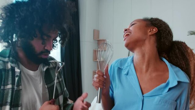 Young happy family African American woman and Arabian man sing songs using kitchen utensils instead of microphone rejoicing in moving to new city or buying apartment on mortgage stands in living room.