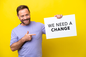 Middle age caucasian man isolated on yellow background holding a placard with text We Need a Change and  pointing it