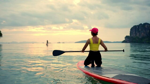 Asian mature woman sitting on paddle board and rowing in the ocean at tropical island at sunset. Wellness woman enjoy outdoor lifestyle and water sports surfing and paddle boarding on summer vacation.