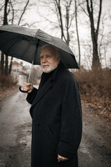 An elderly man in a black coat with an umbrella stands outside in the rain, rainy weather