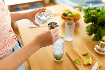 drinks and people concept - close up of woman pouring water from glass bottle to jigger and making lime mojito cocktail at home kitchen