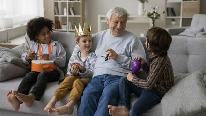 Happy grandson kid and multiethnic friends playing with positive grandpa, using musical toys, crown...