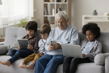 Focused diverse boys and old man sitting on home sofa together, using digital gadgets for online...