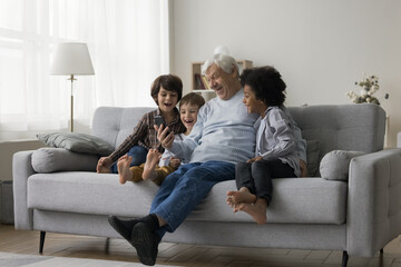 Cheerful happy senior grandfather and multiethnic little grandkids using smartphone for...