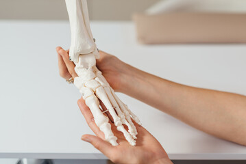 A doctor studies a womans leg structure while holding model of skeletons on a white table indoors....