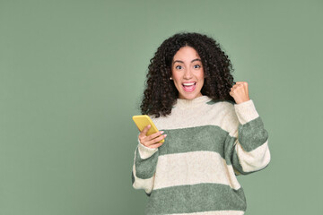 Young happy latin woman winner holding mobile cell phone isolated on green background. Excited...