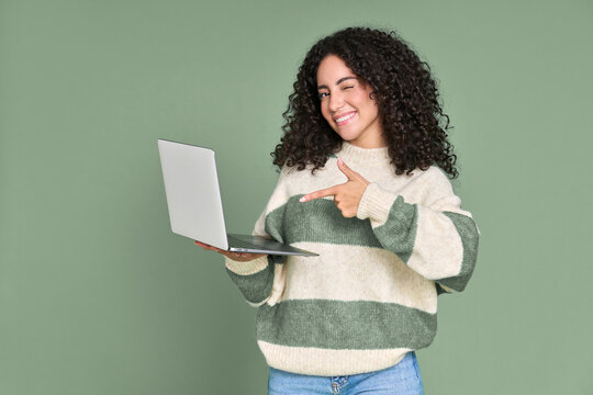 Young happy latin woman winking pointing at laptop isolated on green background. Smiling female model holding computer presenting advertising new trendy website job search or dating service.