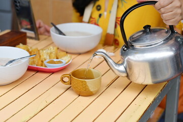 Woman hand pouring hot tea water from aluminum tea pot kettle into brown ceramic mug while eating breakfast in restaurant. The old aluminum pot kettle stove has black handles and fully enclosed lid.
