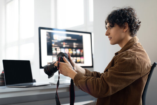 Positive male photographer holding camera while working with photos, looking through taken pictures, working at studio