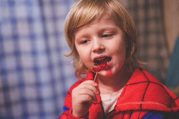 Cute little child in spider man costume eating a red lollipop in form of the number five during his birthday party. Small boy enjoys licking a tasty sugar lollipop. © Татьяна