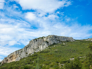 Fototapeta na wymiar Wild landscape of the Alpilles in Provence in France where a rocky outcrop rises from a hill covered in scrubland under a beautiful blue sky adorned with pretty light fluffy white clouds