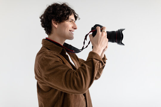Positive male photographer taking photo holding camera near face and smiling, standing over white background, side view