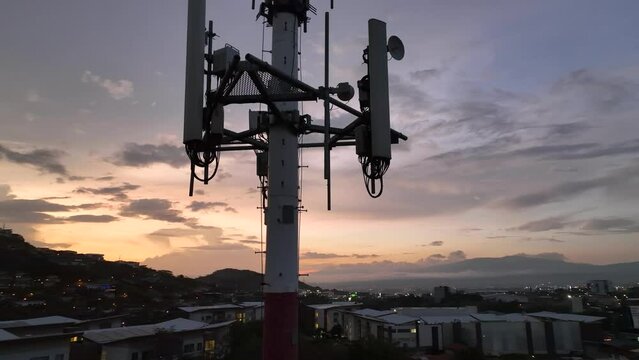 Modern cell tower with cellular network antennas over city at sunset. Close-up drone video rising slowly towards the top.
