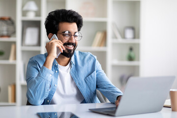 Business Communication. Handsome Indian Male Freelancer Talking On Cellphone And Using Laptop