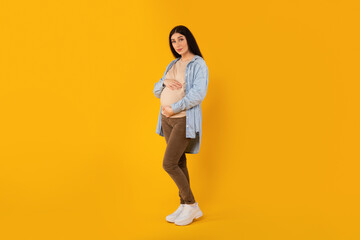 Fototapeta na wymiar Full length portrait of beautiful pregnant lady embracing belly and looking at camera, yellow background, free space