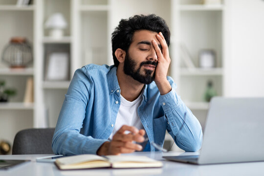 Overworked tired indian male entrepreneur sitting at desk in home office