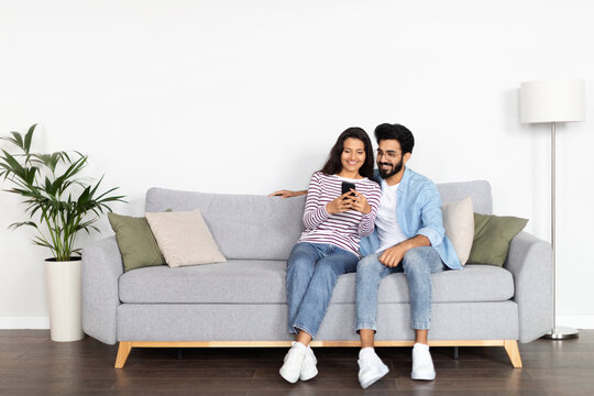 Relaxed multicultural couple sitting on sofa, using smartphone