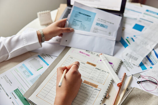 Cropped view of unknown woman sits at desktop with financial papers and bills takes notes of income expenses to daily planner manages expenses and plans future budget reviews reports reads documents