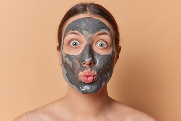 Portrait of shocked brunette woman stares with bugged eyes keeps lips rounded applies facial clay mask for reducing fine lines and pores undergoes beauty procedures isolated over brown background