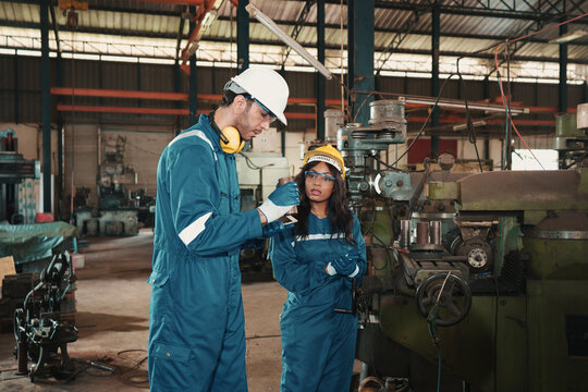 Caucasian factory worker in safety wear is helping African coworker combine a piece of metal part on a machine. Male and female production engineers with responsibility to control metal manufacturing.