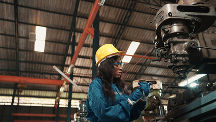Fototapeta premium African factory worker in safety wear is checking a piece of industrial metal while Caucasian coworker help her to combine metal part to machine. Two production engineers are working in metal factory.