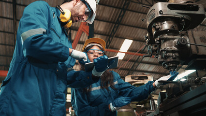 Fototapeta na wymiar African factory worker in safety wear is checking a piece of industrial metal while Caucasian coworker help her to combine metal part to machine. Two production engineers are working in metal factory.