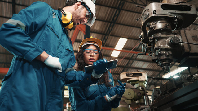 African factory worker in safety wear is checking a piece of industrial metal while Caucasian coworker help her to combine metal part to machine. Two production engineers are working in metal factory.