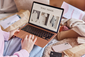Unrecognizable woman uses laptop computer for shopping online searches clothes to buy on resale fashion site surrounded by smartphone credit card and paper packages with clothes makes purchases