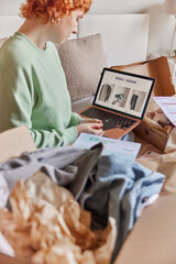 Vertical shot of redhead woman customer makes shopping online holds banking card surfs resale fashion sites on laptop computer surrounded by paper packages with clothes makes order in internet.