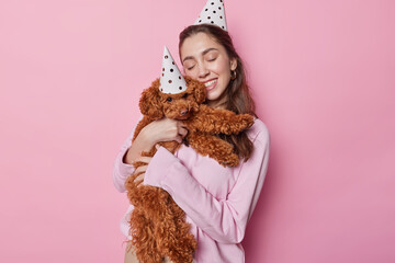 Affectionate brunette woman embraces with love poodle puppy wear party hats celebrate birthday...