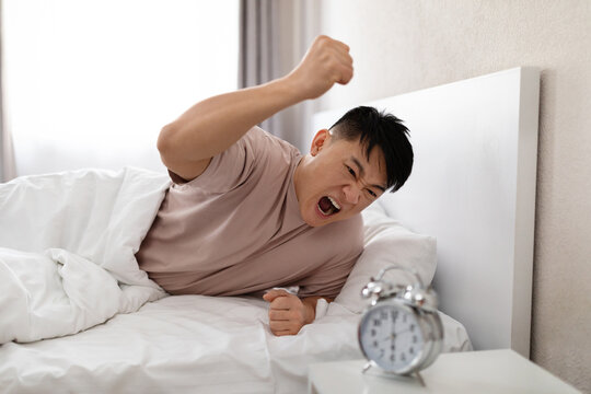 Furious asian man smashing ringing alarm clock, bedroom interior